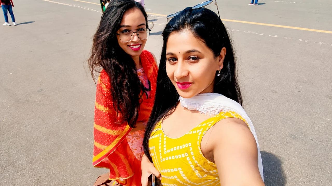 Two school friends taking a selfie outdoors on a sunny day in Delhi, dressed in colorful traditional outfits, smiling as they begin a week of sightseeing, shopping, and summer adventures together.