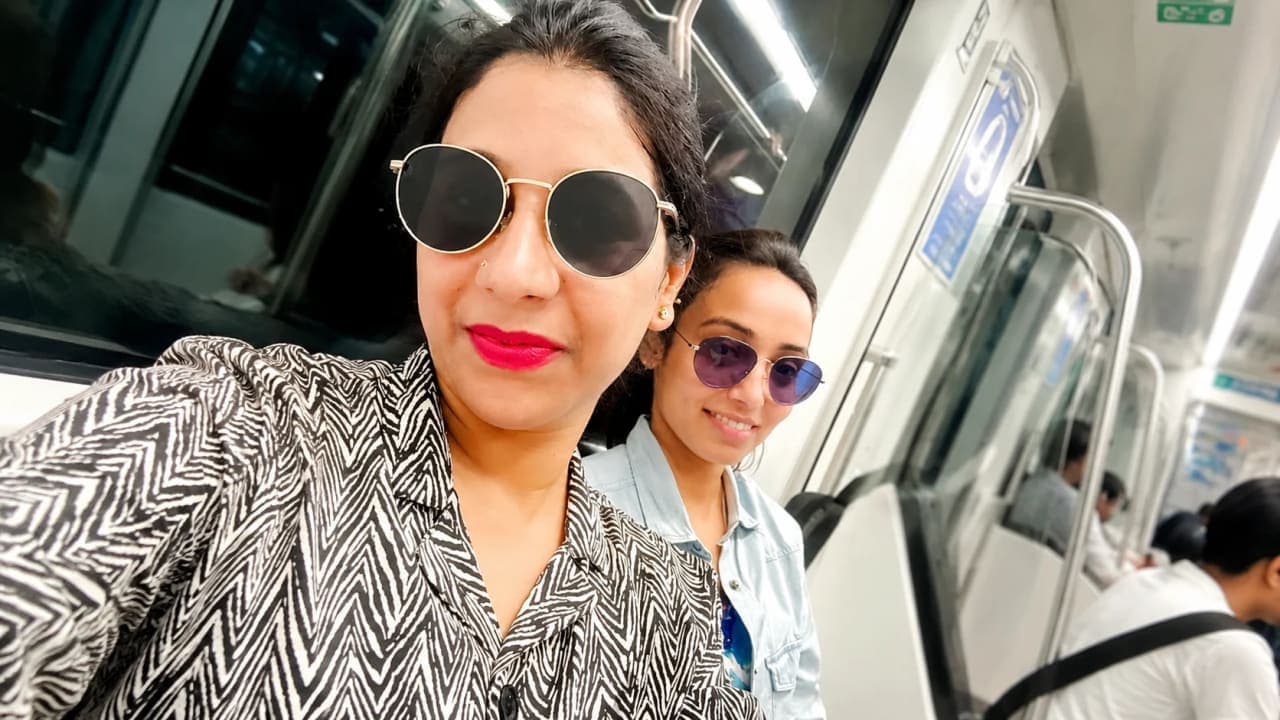 Two friends wearing sunglasses and smiling for a selfie while travelling on the Delhi Metro during a summer trip, with passengers and the metro coach interior visible in the background.