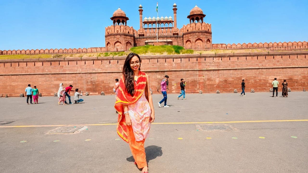 One of the friends posing in front of the historic Red Fort in Delhi on a bright summer afternoon, wearing a colorful traditional outfit while visiting the iconic monument during a week-long trip with a school friend.