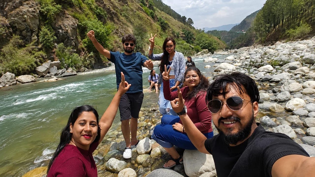 Five friends posing beside a clear mountain river in Himachal Pradesh, enjoying a refreshing break during their journey to Jibhi.