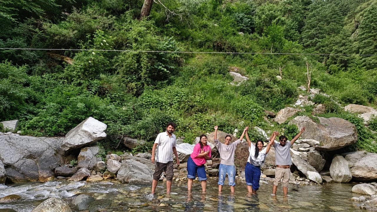 Friends standing and enjoying themselves in a mountain stream near a waterfall in Jibhi, surrounded by rocks, greenery, and flowing water.