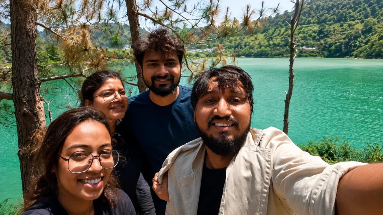 Friends taking a selfie beside a stunning turquoise lake surrounded by green hills during their return journey from Jibhi, stopping briefly to relax and enjoy the scenery.