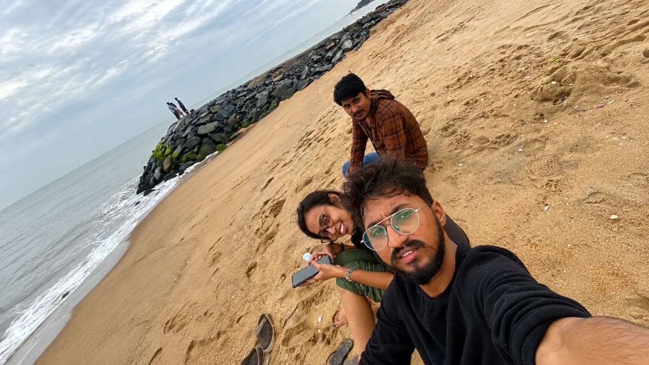 Three friends sitting on a sandy beach in Pondicherry near a rocky shoreline, enjoying a peaceful morning by the sea.