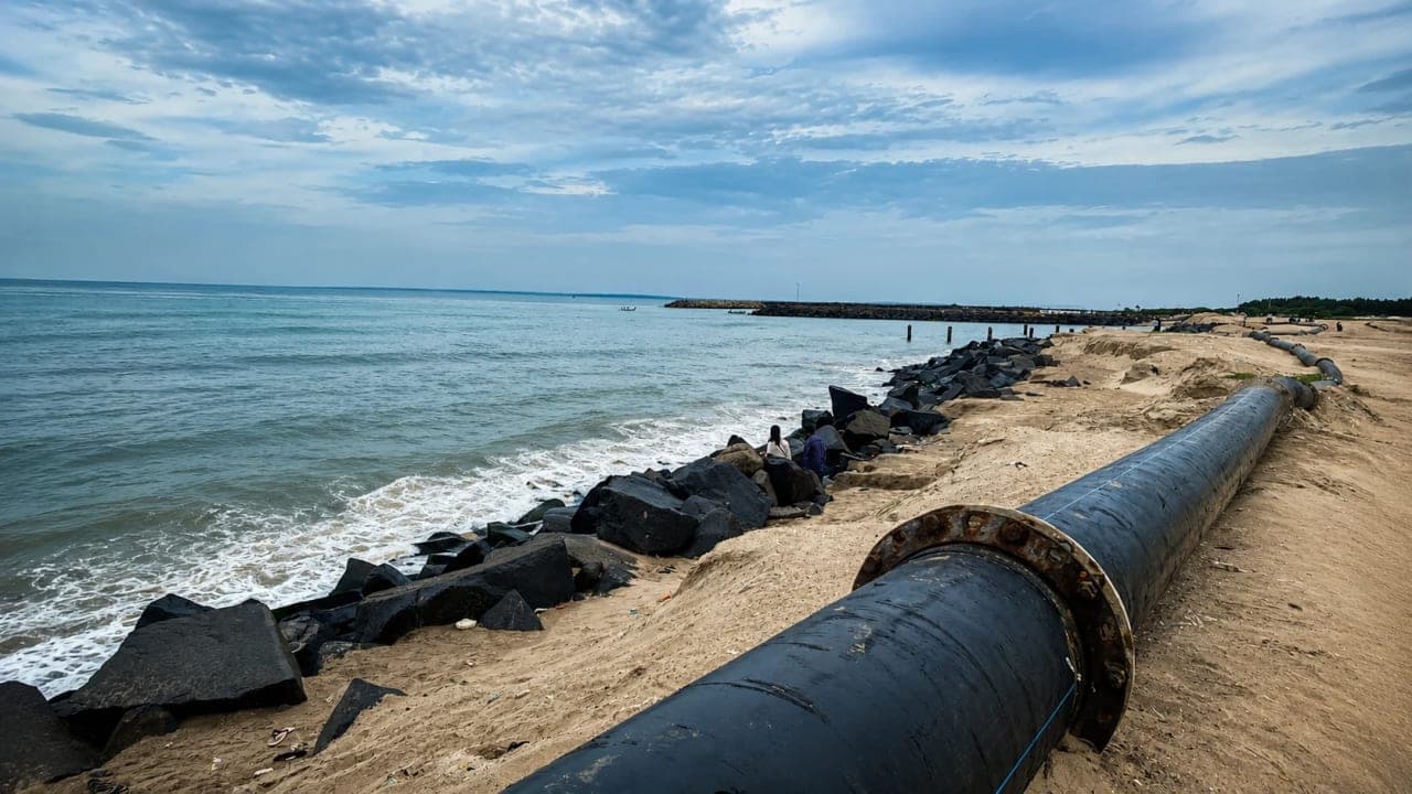 Wide coastal view in Pondicherry featuring rocky shores, calm sea waters, a sandy embankment, and a large pipeline running along the coastline under a cloudy sky.