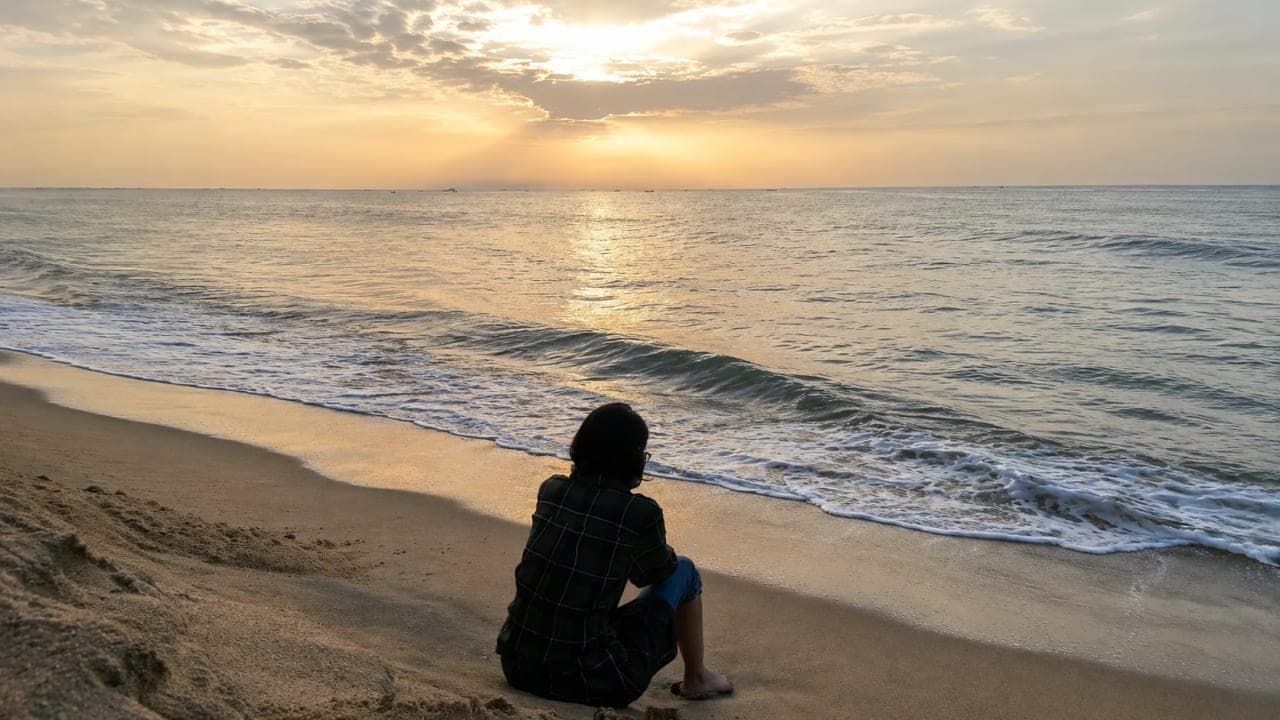 Girl sitting on the sandy beach, facing the sea during a golden sunrise at Auro Beach in Pondicherry.