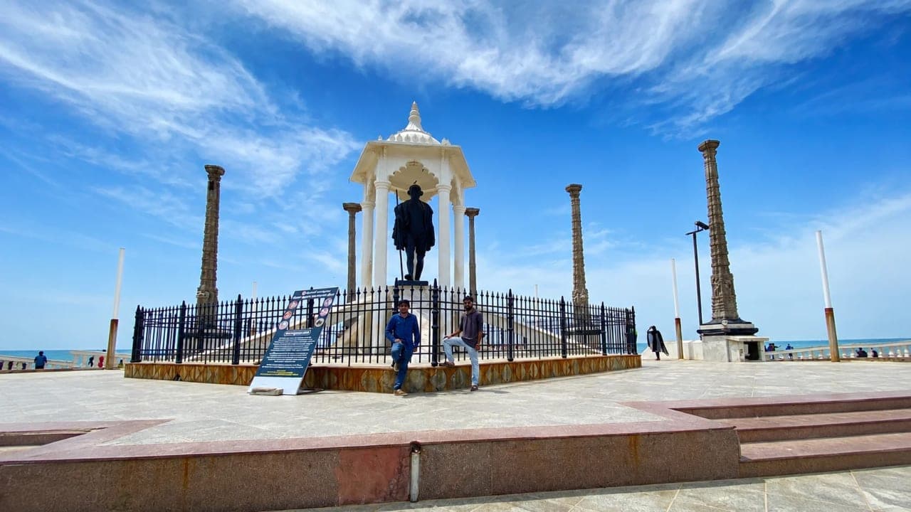 Wide view of the Mahatma Gandhi Statue at Rock Beach in Puducherry with visitors under a bright blue sky.